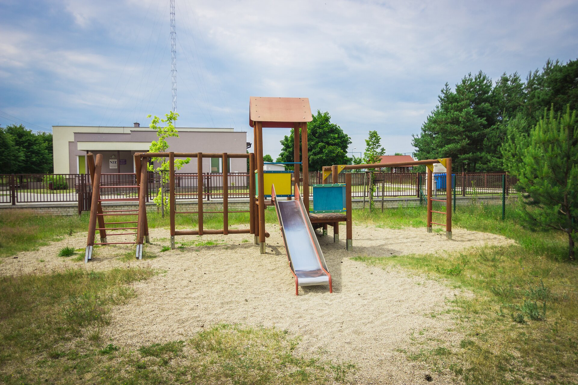 Zdjęcie przedstawia drewniany plac zabaw. Są na nim różne drabinki i niska zjeżdżalnia. W tle widać niski budynek.