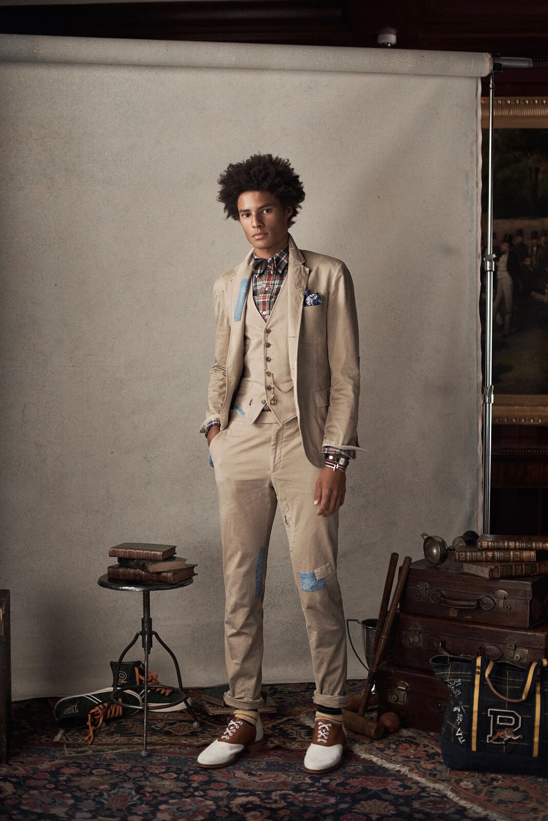 Fotografia przedstawia mężczyznę ubranego w beżowy garnitur z kamizelką, koszulę w czerwono‑beżową drobną kratę, biało‑brązowe garniturowe buty oraz dwie bransolety na lewej ręce. Garnitur ma niebieskie oraz jasnokremowe przeszycia przypominające łatki, a powyżej butów widoczne są czarno‑żółte skarpety. Włosy mężczyzny to średniej długości uniesione afro. Stoi on tle szarego materiału, obok niego widoczny jest mały stolik z książkami oraz skrzynie, torba, kij do gry w polo i para trampek za kostkę. Na podłodze położony jest dywan w nieregularne wzory, a na widocznym fragmencie ściany można zauważyć obraz ukazujący elegancko ubranych mężczyzn w kapeluszach. 