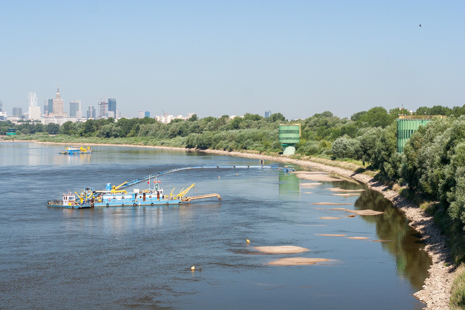 Zdjęcie przedstawia rzekę. Na wodzie znajduje się łódź transportowa. Na jej prawym brzegu rosną drzewa. W oddali znajduje się centrum miasta i należące do niego wieżowce. 