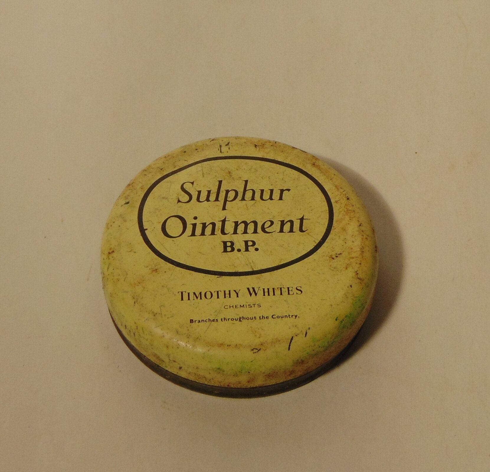 Zdjęcie przedstawiające opakowanie maści siarkowej. Opakowanie w kształcie koła płaskie z zaokrąglonymi brzegami. Na opakowaniu widniej napis Sulphur ointment B. P. Timothy Whites.