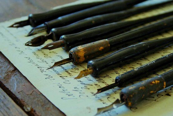 Zdjęcie przedstawia czarne, drewniane obsadki ze stalówkami używane dawniej do pisania atramentem lub tuszem na papierze. Osiem obsadek leży równo obok siebie na kartce zapisanej odręcznym pismem. Widać, że są stare, wielokrotnie używane, z dwóch odchodzi czarna farba. Każda z nich ma założoną stalówkę o innym kształcie.