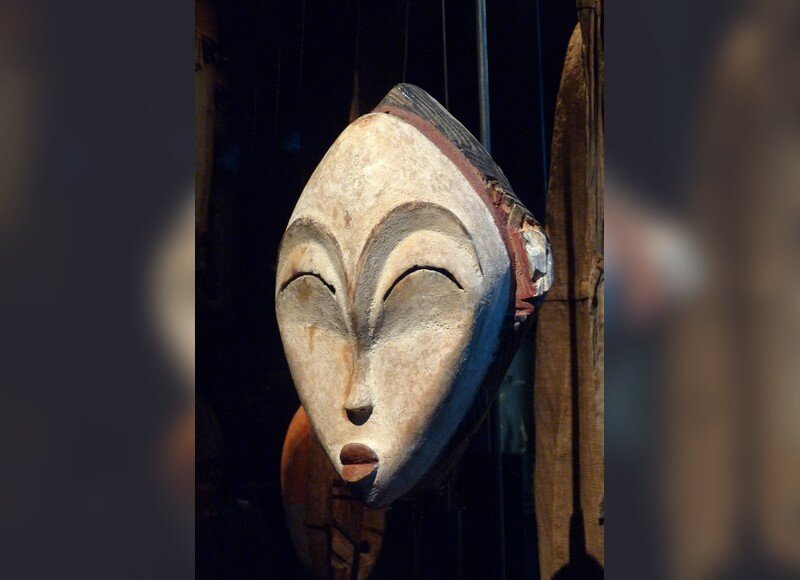 Zdjęcie przedstawia drewnianą maskę. Maska ma owalny kształt, ma bardzo duże, łukowate brwi i zamknięte powieki.