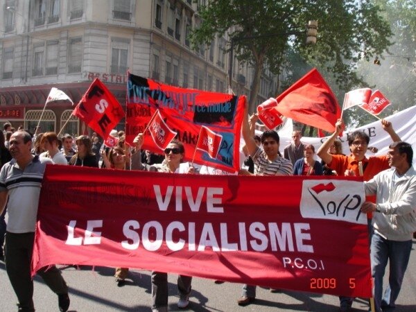 Zdjęcie przedstawia pochód ludzi ulicami miasta. Na pierwszym planie dwóch mężczyzn trzyma duży, czerwony transparent z napisem Vive le socialisme. Za nim idą inni ludzie, niosąc kolejne, zwykle czerwone transparenty z hasłami. W tle jest kilkupiętrowy miejski budynek. Zdjęcie wykonano 5 maja 2009 roku. 