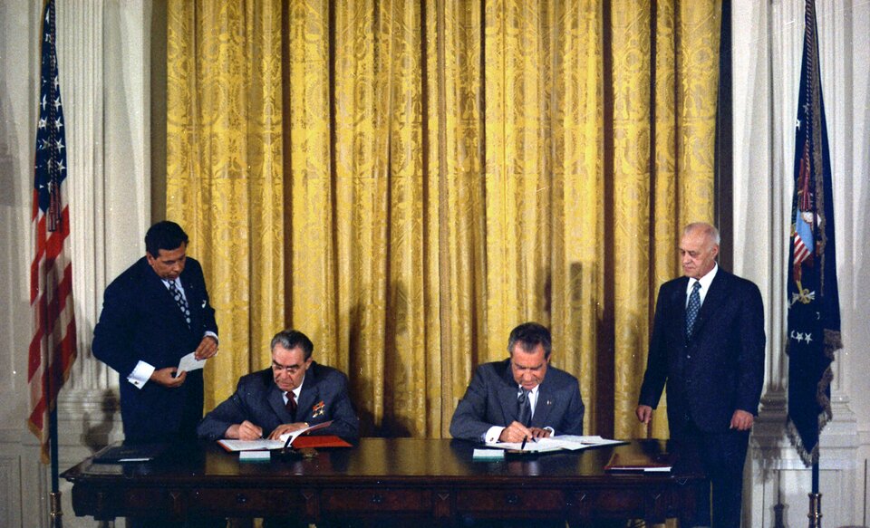 Zdjęcie przedstawia dwóch mężczyzn podpisujących dokumenty przy prostokątnym stole z ciemnego drewna. Za podpisującymi wisi złota kotara. Po bokach stoją dwie flagi, po lewej flaga Stanów Zjednoczonych.