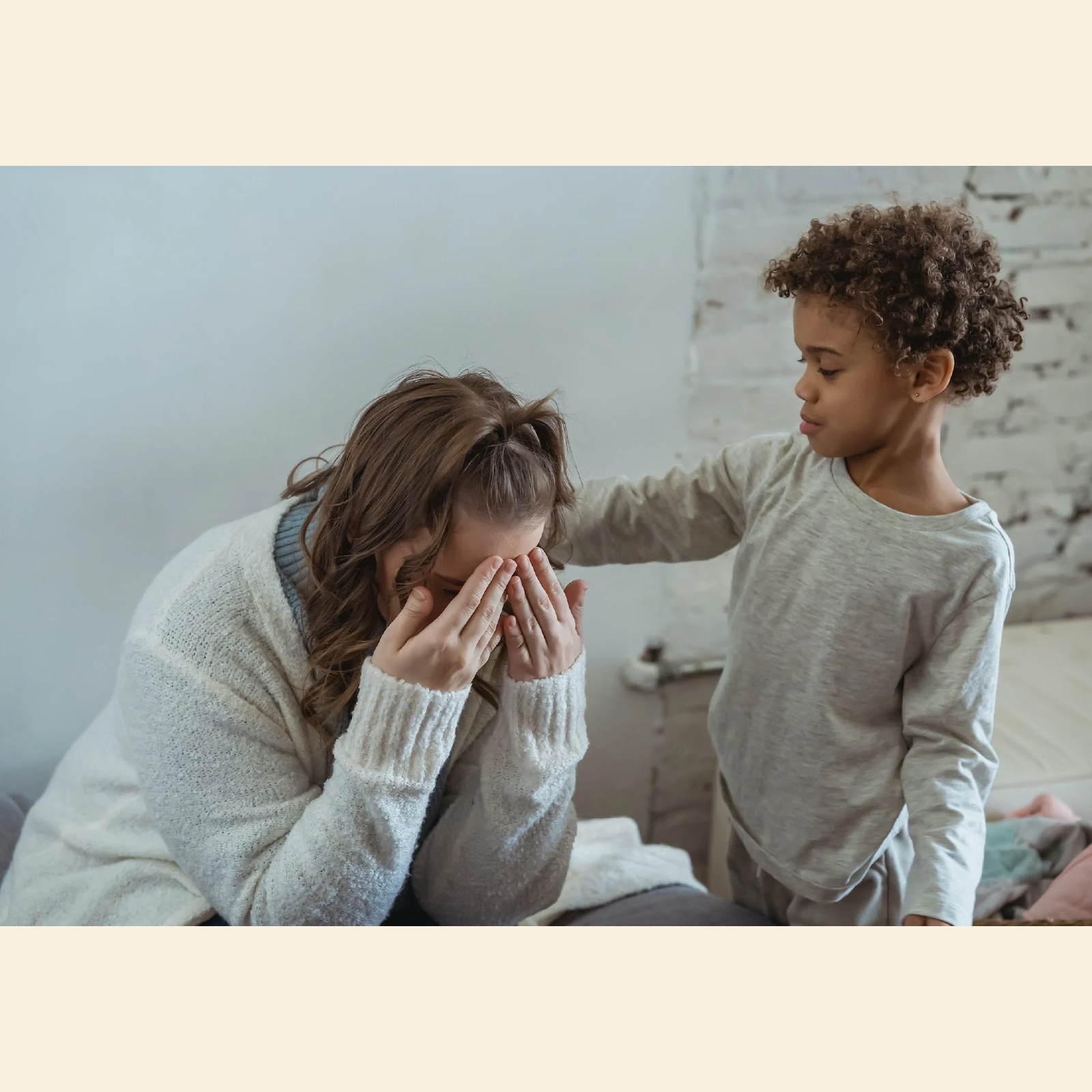 Ilustracja przedstawia siedzącą kobietę zakrywającą twarz dłońmi. Kobieta ma biały sweter sięgający za nadgarstki. Obok stoi chłopiec w wieku wczesnoszkolnym z włosami kręcącymi się w loczki. Chłopiec opiera rękę na ramieniu kobiety okazującej wsparcie.