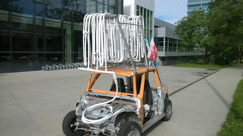 Zdjęcie przedstawia pojazd zbudowany na bazie szkieletu Fiata sto dwadzieścia sześć pe ze skomplikowanym układem białych rurek na dachu. Gruby przewód od tych rurek biegnie do układu znajdującego się gdzieś pod srebrną częścią obudowy z ocynkowanej stali lub aluminium. Pojazd ma dwa miejsca siedzące, jest pozbawiony szyb i drzwi. Stoi na betonowej płycie przed oszklonym budynkiem uczelni.