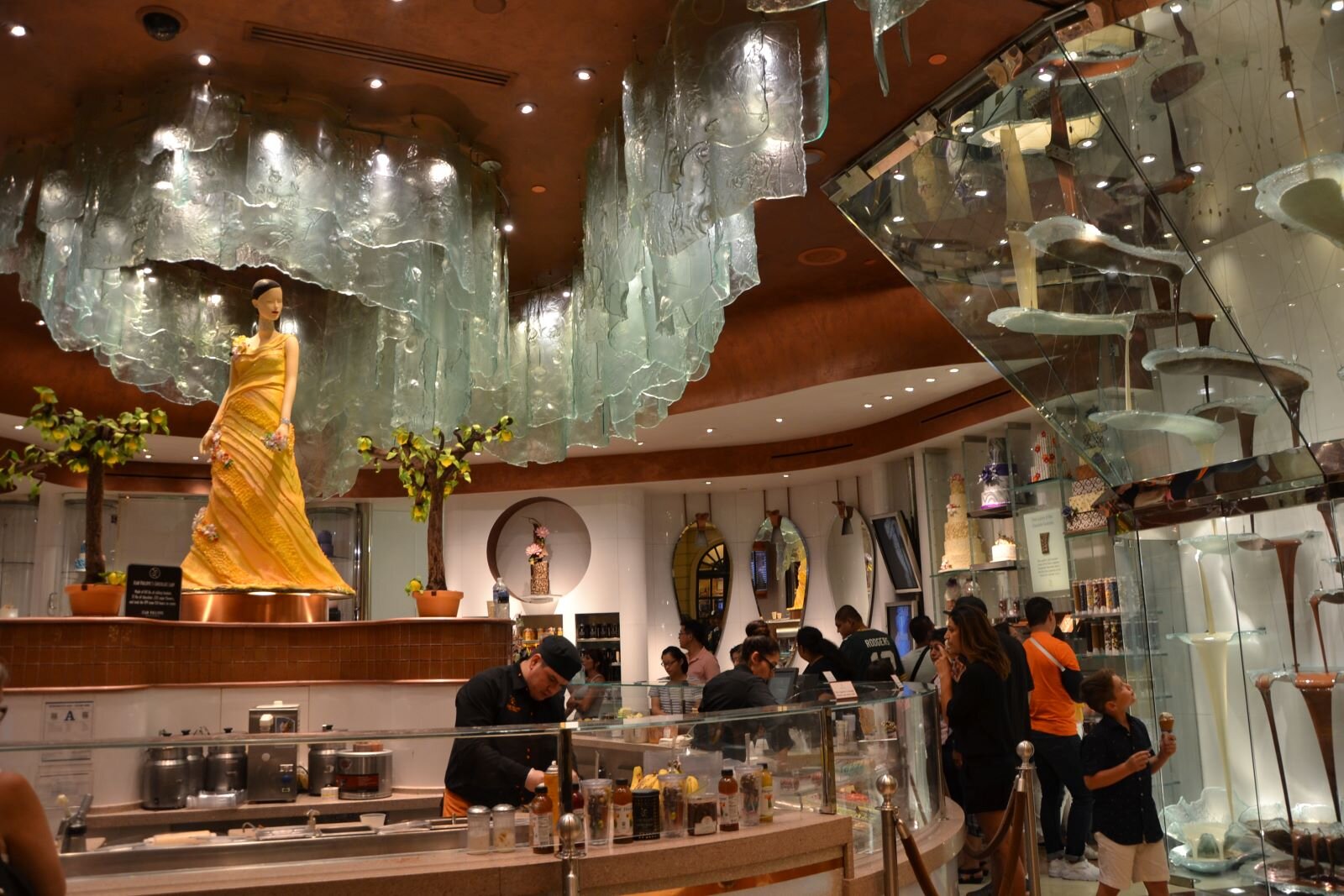 Ilustracja przedstawia wnętrze sklepu z czekoladą, w którym znajduje się wiele klientów. Wnętrze jest bogato dekorowane: na suficie jest ogromnych rozmiarów szklany zwisający żyrandol, który zajmuje większość sufitu - składa się z jednakowych elementów gęsto ułożonych i skierowanych w dół. Na ścianach znajdują się ogromne tafle lustrzane na tle których umieszczone są fantazyjne eskpozycje z czekolady. Elementem najbardziej przyciągającym uwagę jest ogromny posąg kobiety w pomarańczowej sukni, stojący wysoko na cokole sięgającym ponad głowy stojących ludzi. Dzięki takiemu umieszczeniu głowa posągu sięga szklanych elementów żyrandola.