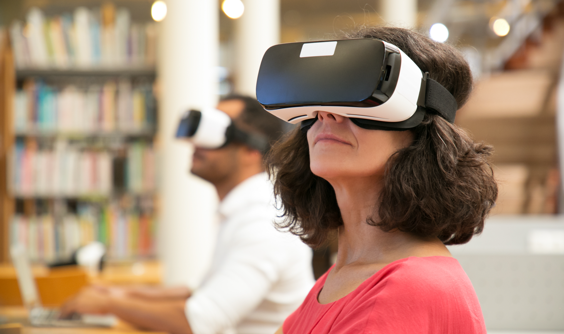 Zdjęcie przedstawia dwie osoby siedzące w bibliotece przy biurkach w goglach elektronicznych.