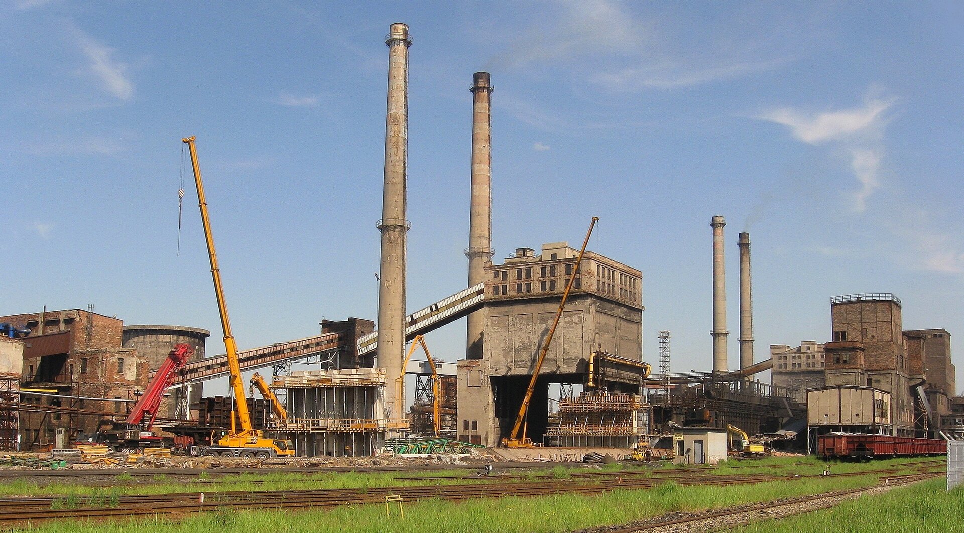 Zdjęcie przedstawia fabryczne zabudowania z kilkoma wysokimi kominami. W pobliżu fabryki są tory kolejowe.  