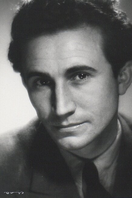 Zdjęcie przedstawia twarz młodego mężczyzny. Jest ona pociągła, nos wydatny, brwi wyraziste, oczy głęboko osadzone. Mężczyzna ma krótkie, falujące, zaczesane ku górze włosy.