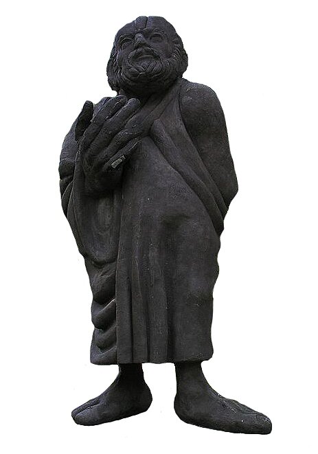 Zdjęcie przedstawia rzeźbę starszego mężczyzny. Jego gołe stopy i prawa dłoń, którą podnosi na wysokości piersi, są nieproporcjonalnie duże. Twarz okolona jest bujnym zarostem. Postać jest odziana w długą, luźno zwisającą szatę.