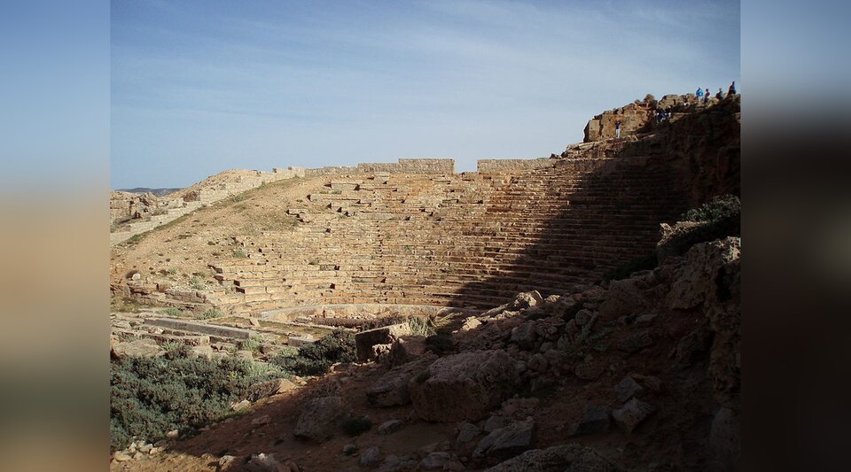Zdjęcie przedstawia ruiny starożytnego teatru Dionizosa. Ukazana jest widownia usytuowana na niewielkim wzniesieniu. Siedzenia dla widzów są poniszczone.