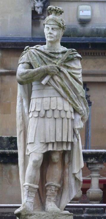 Fotografia przedstawia męski posąg, wykonany z jasnego kamienia. Mężczyzna ubrany jest w krótką zbroję, płaszcz oraz wysoko wiązane  sandały. Na głowie ma ozdobny hełm. W prawej ręce trzyma zwinięty papirus z małym pióropuszem.  W tle znajduje się fragment szarego współczesnego budynku.