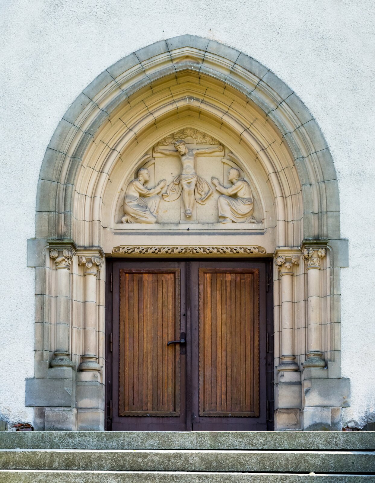 Zdjęcie przedstawia prostokątne, dwuskrzydłowe, drewniane drzwi. Dookoła nich ozdobny portal, zakończony ostrym łukiem. Nad drzwiami znajduje się trójkątna wnęka, w której umieszczono rzeźbę Jezusa na krzyżu z dwoma postaciami ze skrzydłami, klęczącymi po obu jego stronach.