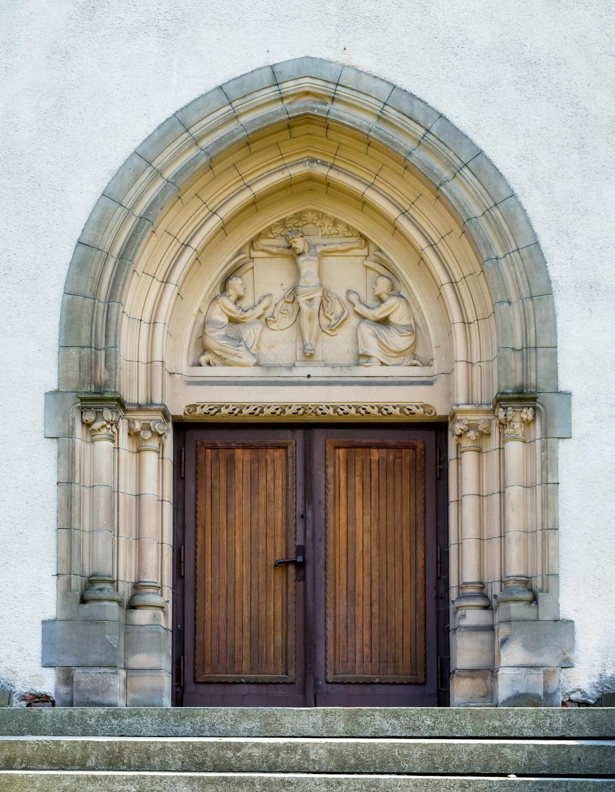 Zdjęcie przedstawia prostokątne, dwuskrzydłowe, drewniane drzwi. Dookoła nich ozdobny portal, zakończony ostrym łukiem. Nad drzwiami znajduje się trójkątna wnęka, w której umieszczono rzeźbę Jezusa na krzyżu z dwoma postaciami ze skrzydłami, klęczącymi po obu jego stronach.