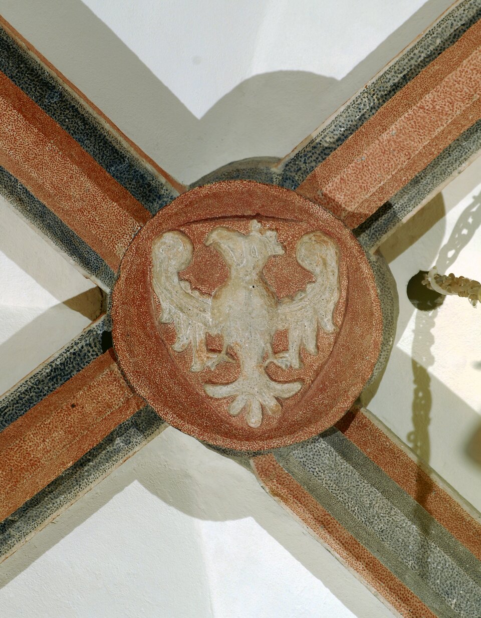 Ilustracja przedstawia zwornik z wizerunkiem orła białego na czerwonym tle. Od zwornika wychodzą cztery odnóża - trzy czerwone z czarnymi detalami oraz jedno szare z czerwonym detalem.