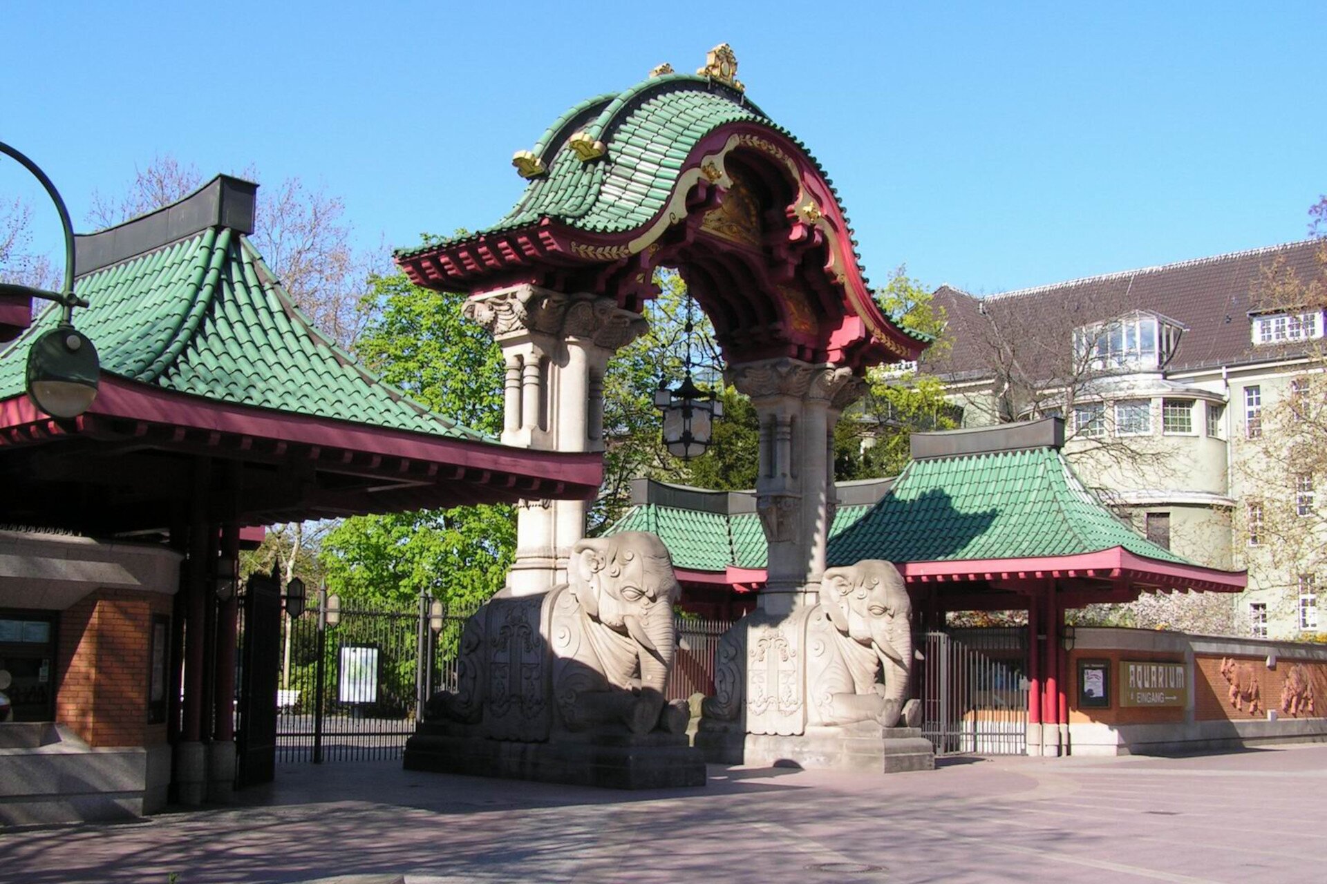 Ilustracja przedstawia wejście do ogrodu zoologicznego. Przy bramie ze spadzistym dachem stoją dwie rzeźby przedstawiające słonie.