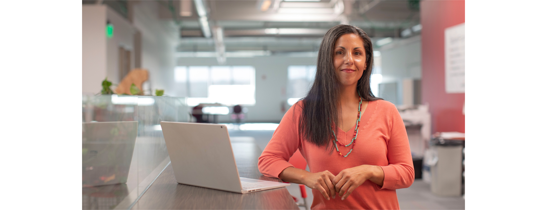Zdjęcie przedstawia kobietę z długimi włosami w pomieszczeniu. Obok kobiety na blacie stoi otwarty laptop.