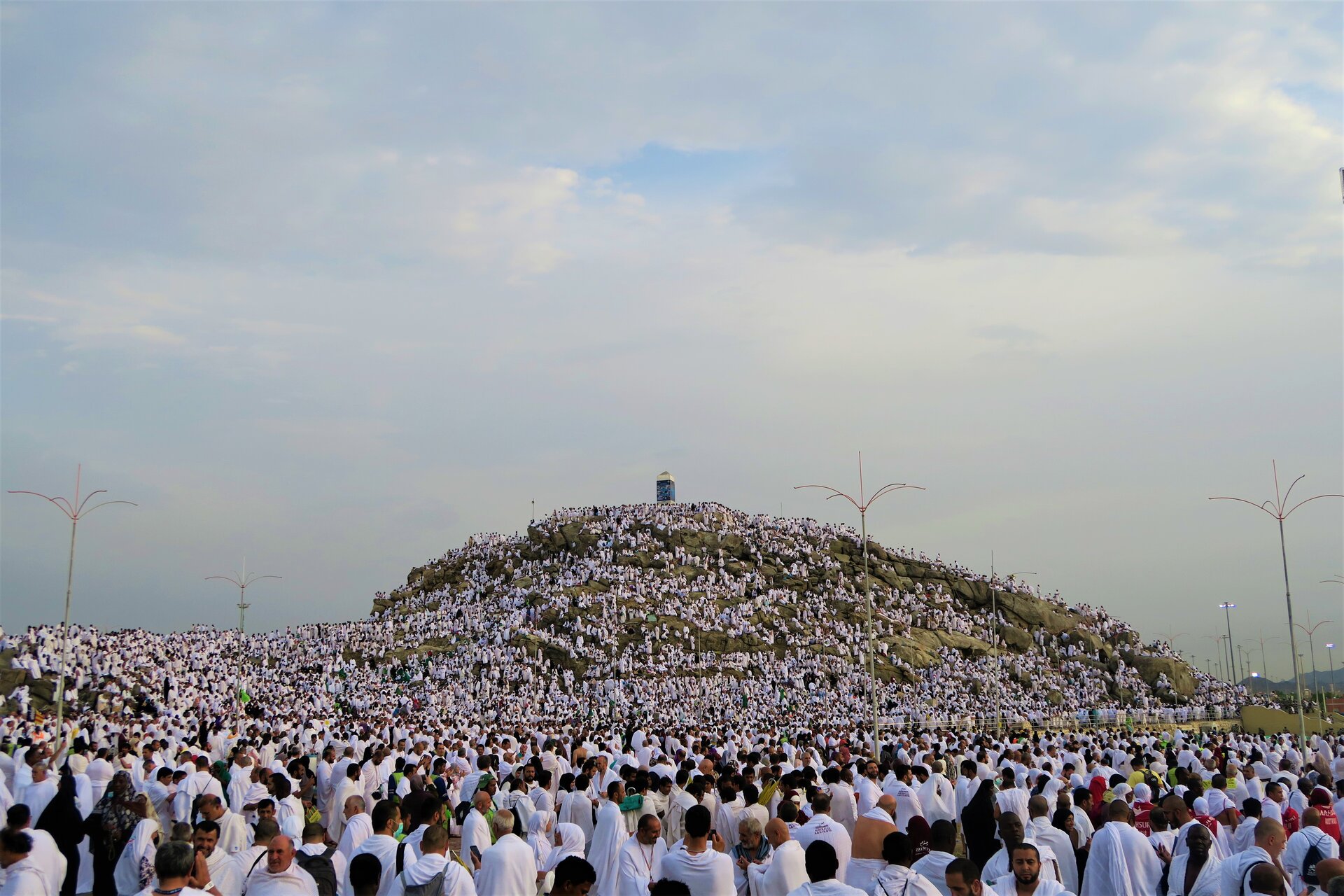 Zdjęcie przedstawia niewysokie wzgórze, na którym znajduje się tłum ludzi. Pielgrzymi ubrani są w białe szaty. Na szczycie wzgórza stoi obelisk.  