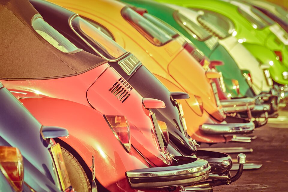 Zdjęcie przedstawia rząd klasycznych samochodów w żywych kolorach – czerwonym, niebieskim, czarnym, żółtym, zielonym i innych – ustawionych w linii na parkingu. Są to kultowe Volkswageny Garbusy, rozpoznawalne po zaokrąglonych kształtach i charakterystycznej tylnej części. Zdjęcie jest wykonane pod kątem, koncentrując się na detalach tylnych błotników, reflektorów i chromowanych zderzaków.