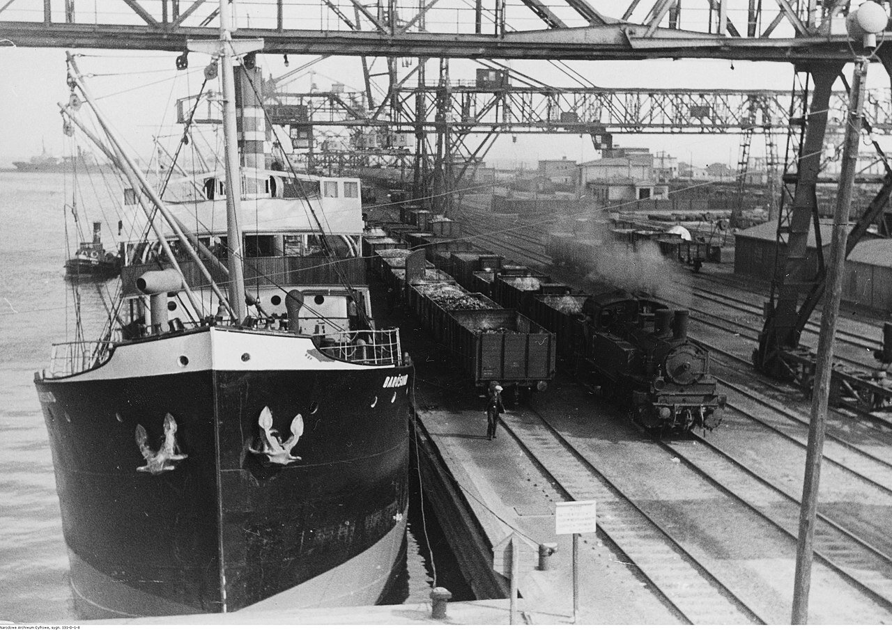 Zdjęcie przedstawia statek, za nim widoczna jest kilka razy mniejsza łódź. Po prawej stronie od statku, już na lądzie, kilka torów kolejowych. Na jednym z nich stoi pociąg towarowy a na drugim same kontenery szynowe, bez lokomotywy. Przewożony jest w nich jakiś surowiec. Na górze zdjęcia widoczne są rusztowania, w tle budynki stoczniowe. Fotografia czarno‑biała.