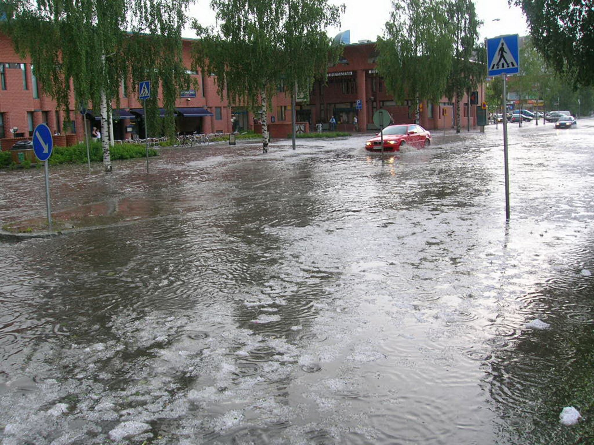 Na zdjęciu zalana ulica w mieście po przejściu ulewy. Woda ma dość wysoki poziom, sięga niemal do połowy kół samochodów poruszających się po drodze.