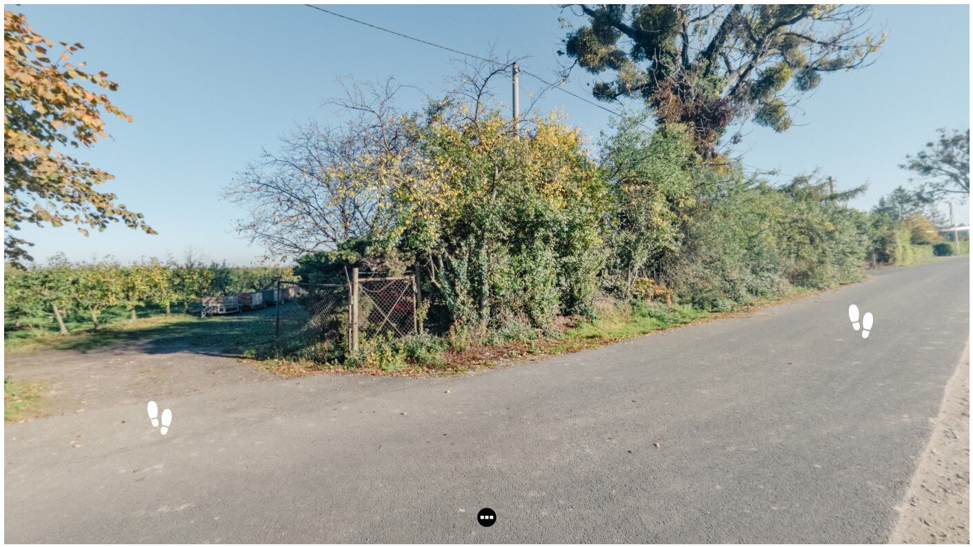 Plansza z piktogramami stóp umożliwiającymi przejście między obszarami sadu. Zdjęcie przedstawia drogę asfaltową, przy której rosną drzewa i krzewy. Na drodze w dwóch miejscach znajdują się białe ślady stóp.