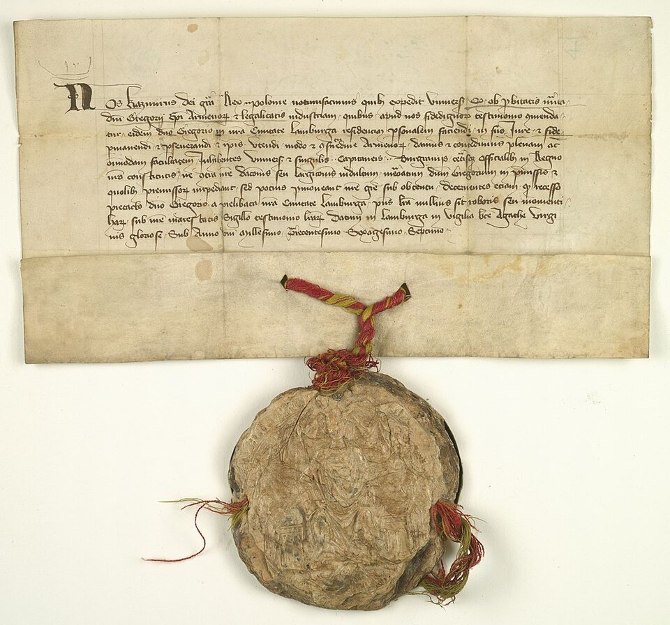 Zdjęcie przedstawia dokument zapisany pismem odręcznym w języku łacińskim. Pismo jest drobne, kaligrafowane z fantazyjnymi zawijasami. U dołu dokumentu znajduje się okrągła pieczęć zawieszona na sznurze.