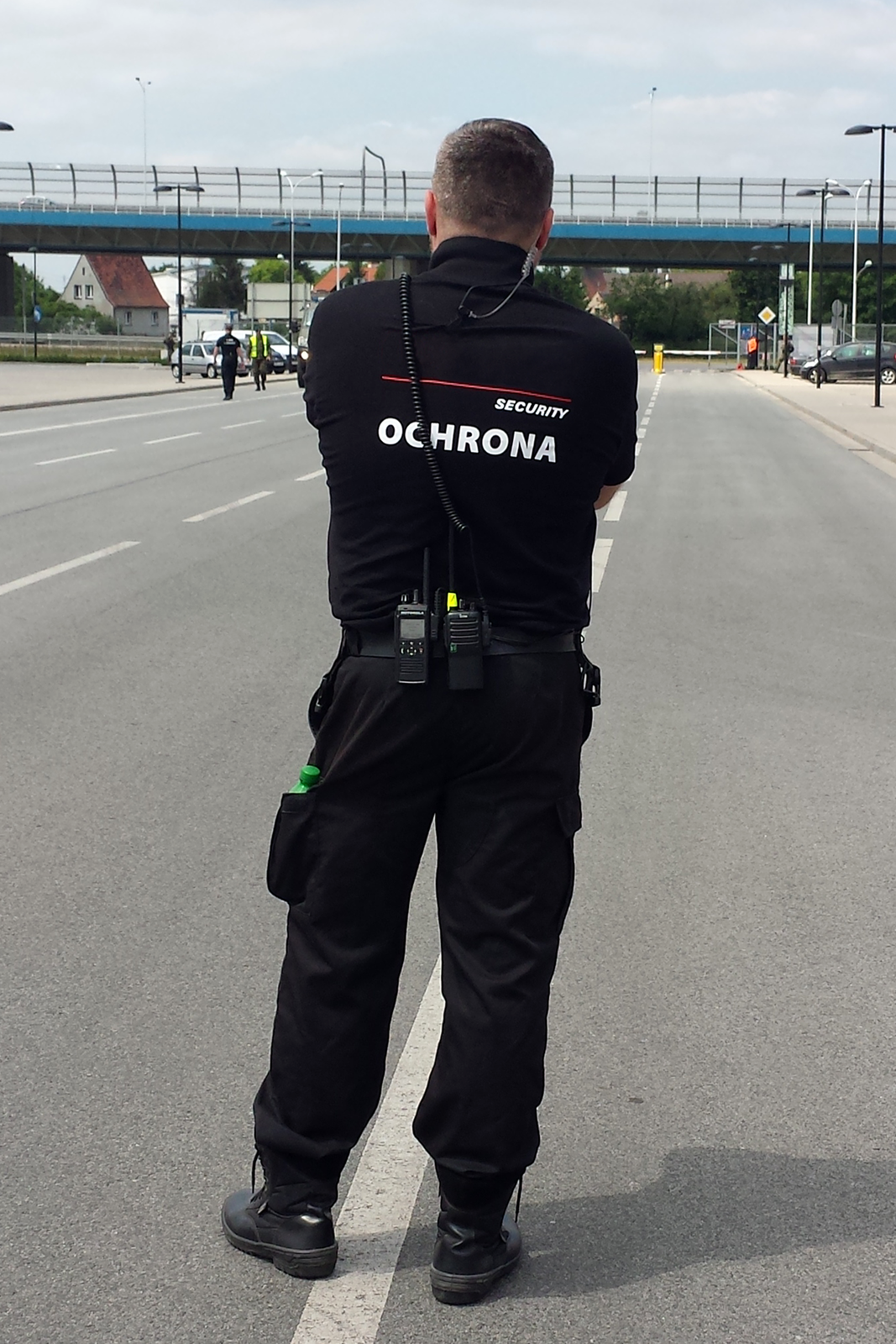 Kolorowe zdjęcie przedstawia stojącego tyłem pracownika firmy ochroniarskiej. Słoneczny dzień. Pracownik stoi na środku czteropasmowej ulicy. Ubrany w czarny mundur firmowy. Na pasku, poniżej pleców, przytwierdzone telefony i nadajnik. Na plecach biały napis: security ochrona. Mężczyzna obserwuje okolicę. W głębi zdjęcia wiadukt nad ulicą.