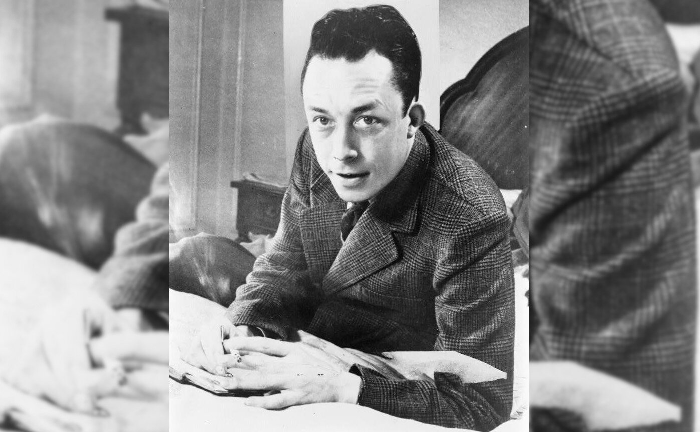 Zdjęcie przedstawia dojrzałego mężczyznę. Patrzy w stronę widza. Jego twarz jest owalna, nos duży, prosty, brwi wyraziste. Mężczyzna ma krótkie włosy zaczesane ku górze. Mężczyzna siedzi, ma ręce położone na otwartej książce. Jest ubrany w kraciastą marynarkę.