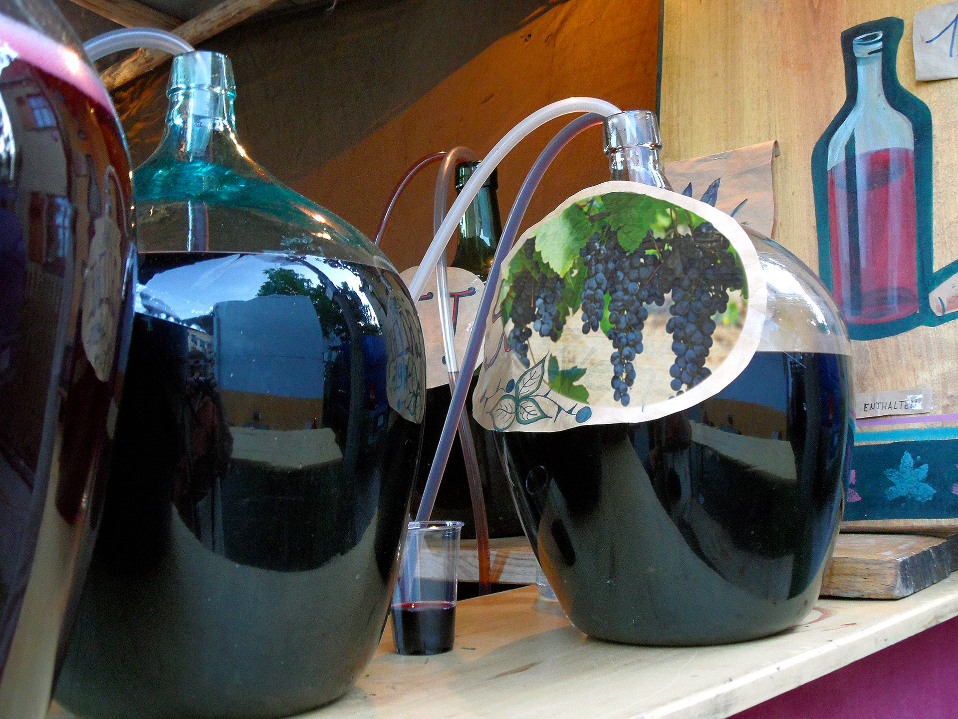 Zdjęcie przedstawia dwa galony na wino. Wino powstaje w wyniku fermentacji owoców. Pędzone jest z ciemnych winogron. Z balonu wystaje giętka rurka, którą spuszcza się małą ilość powstałego wina do szklanki