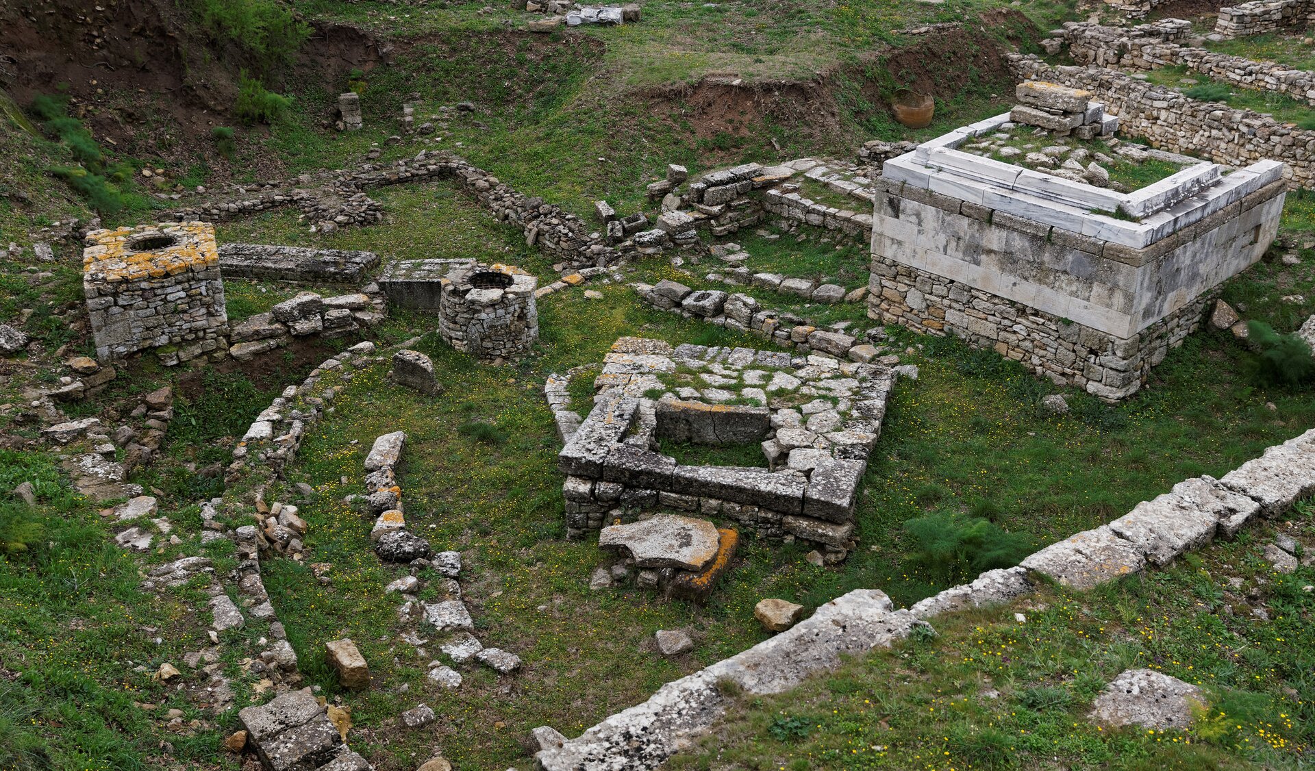 Zorientowana poziomo fotografia barwna przedstawiająca nieco z góry fragment ruin starożytnej Troi znajdujących się obecnie na terytorium Turcji. Widać ułożone obok siebie kamienne bloki, wystające spod zielonej trawy, które tworzą fundamenty budowli. W prawym widoczne są około metrowe fundamenty kwadratowej budowli ułożone z płaskich kamieni. 
