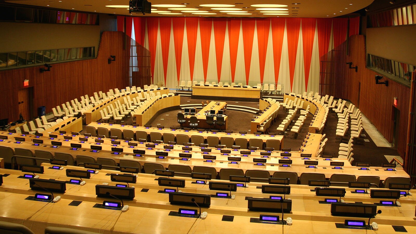 Zdjęcie przedstawia dużą salę obrad, w której w przedniej części widoczne są fotele ustawione po łuku. Pośrodku widoczny jest stół w kształcie litery T, na którym stoją monitory komputerów. W tle widoczny jest duży, prostokątny stół prezydialny z rzędem foteli. Na pierwszym planie widoczne są jasnobrązowe blaty, na których znajdują się małe mikrofony, wyświetlacze oraz przyciski do głosowania. Na ścianie, za stołem prezydialnym, widoczna jest tkanina we wzór naprzemiennie ułożonych czerwonych i jasnożółtych trójkątów.