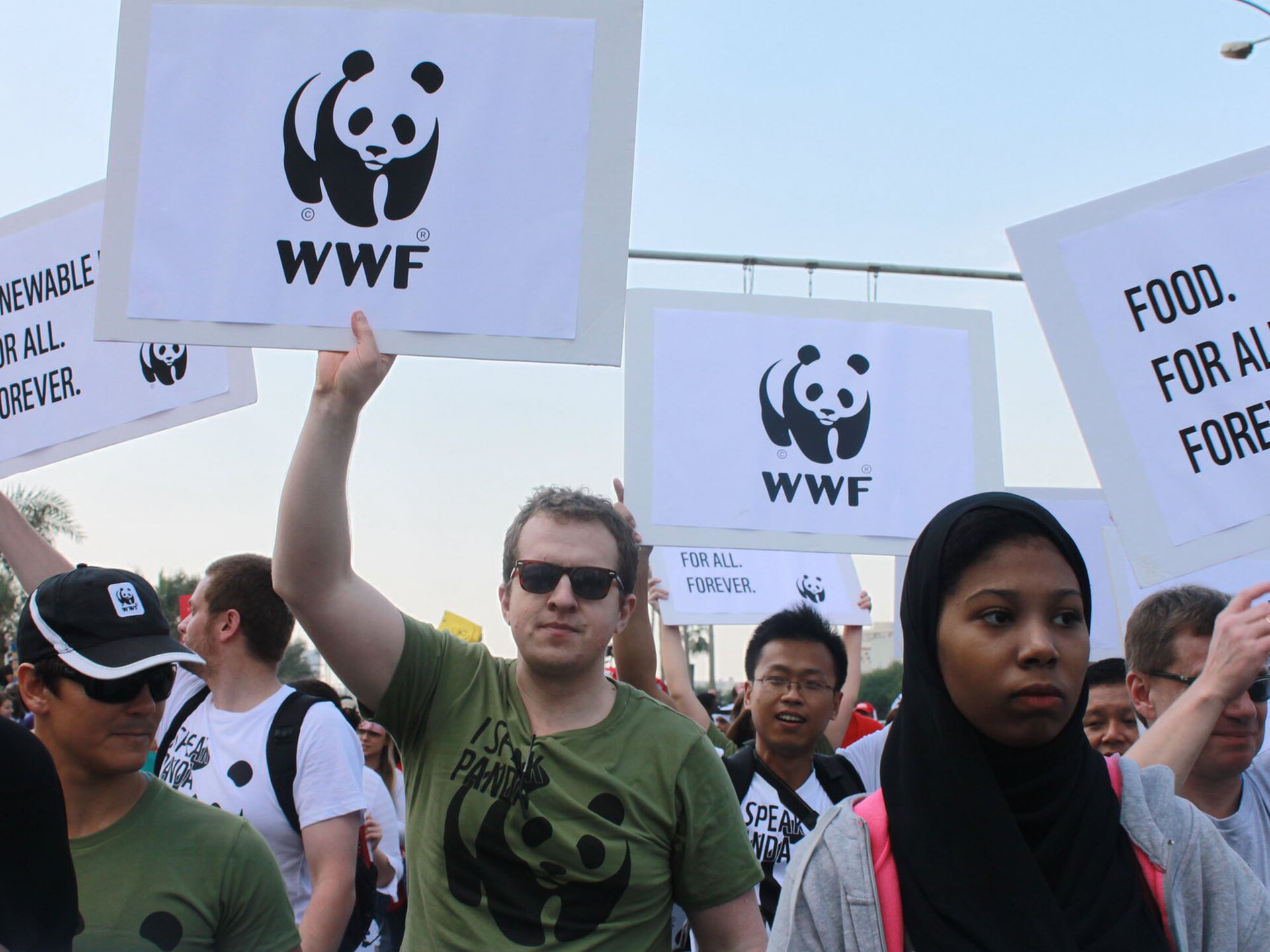 Zdjęcie przedstawia manifestację osób wspierających organizację WWF. Widoczne są idące w marszu osoby, które trzymają transparenty z logo WWF, które złożone jest z czarno-białej pandy, pod którą znajdują się litery WWF. Na innych transparentach widoczne są fragmenty haseł w języku angielskim: FOOD. FOR ALL. FOREVER.