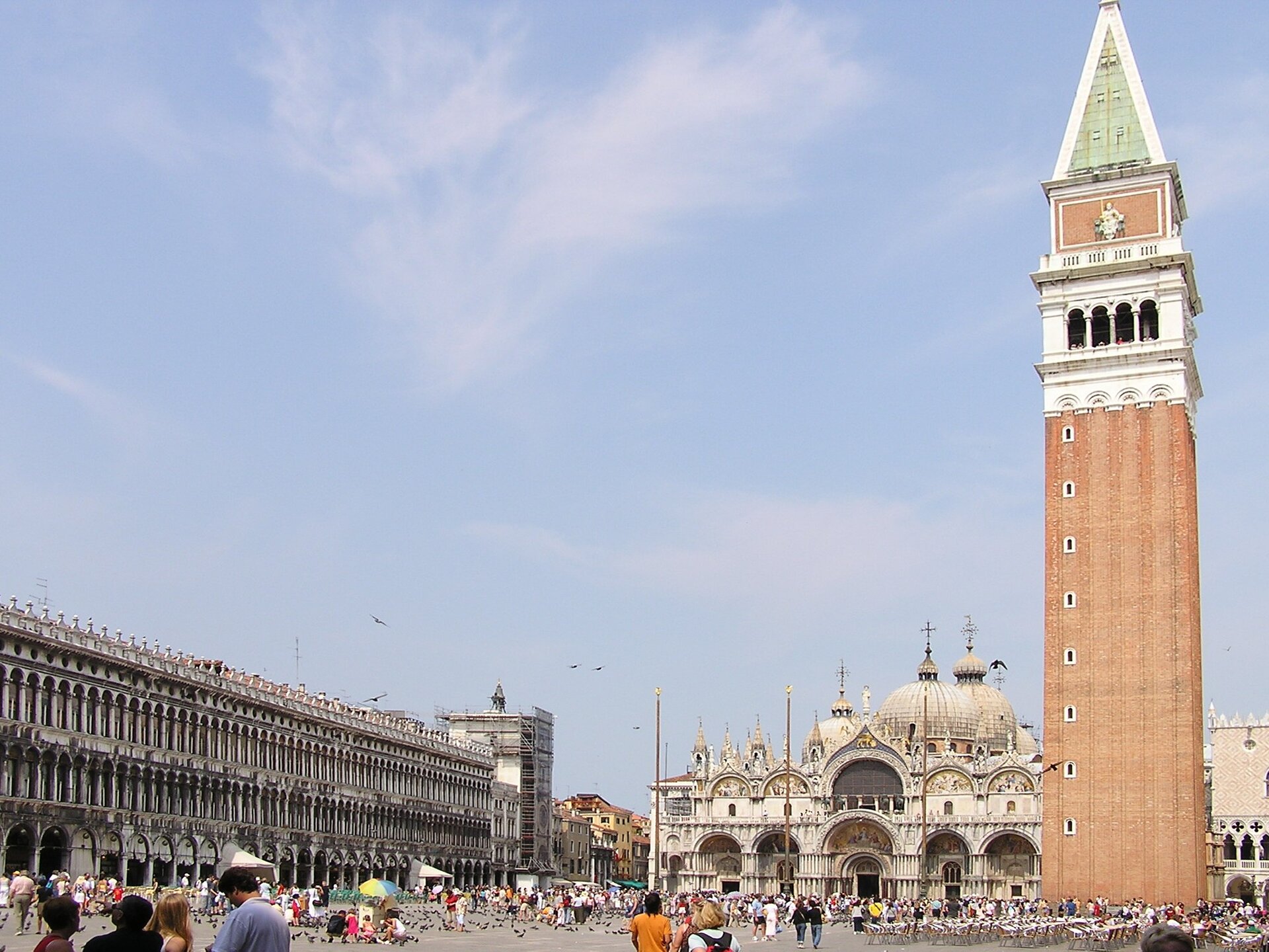 Plac Świętego Marka w Wenecji. Na zdjęciu rozległy plac z monumentalnymi budynkami, obszerna bazylika ze zdobioną fasadą, licznymi kopułami, wieżami, z prawej strony – wysoka ceglana dzwonnica. Na placu duża liczba turystów. W tle bezchmurne, jasnoniebieskie niebo.