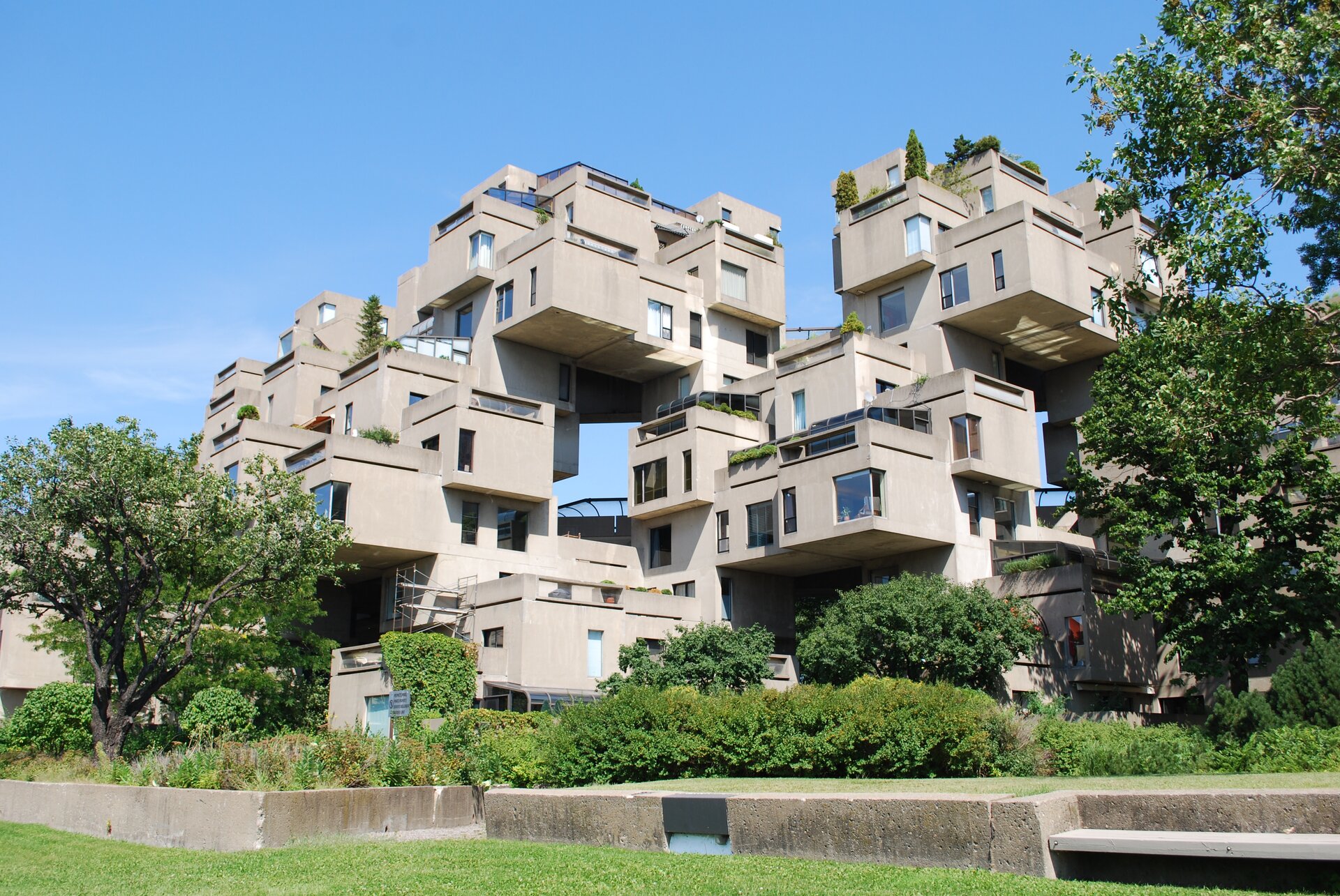 Ilustracja przedstawia fragment Habitat 67. Jest to kompleks mieszkalny w Montrealu w Kanadzie, wzniesiony na wystawę światową w 1967 roku, gdzie pełnił rolę jednego z pawilonów. Dookoła budowli znajdują się liczne drzewa oraz krzewy.