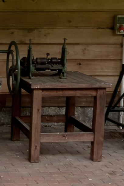 Zdjęcie przedstawia taśmiarkę ręczną stojąca na zewnątrz budynku. Urządzenie stoi na drewnianym stole i posiada duże koło zamachowe. 
