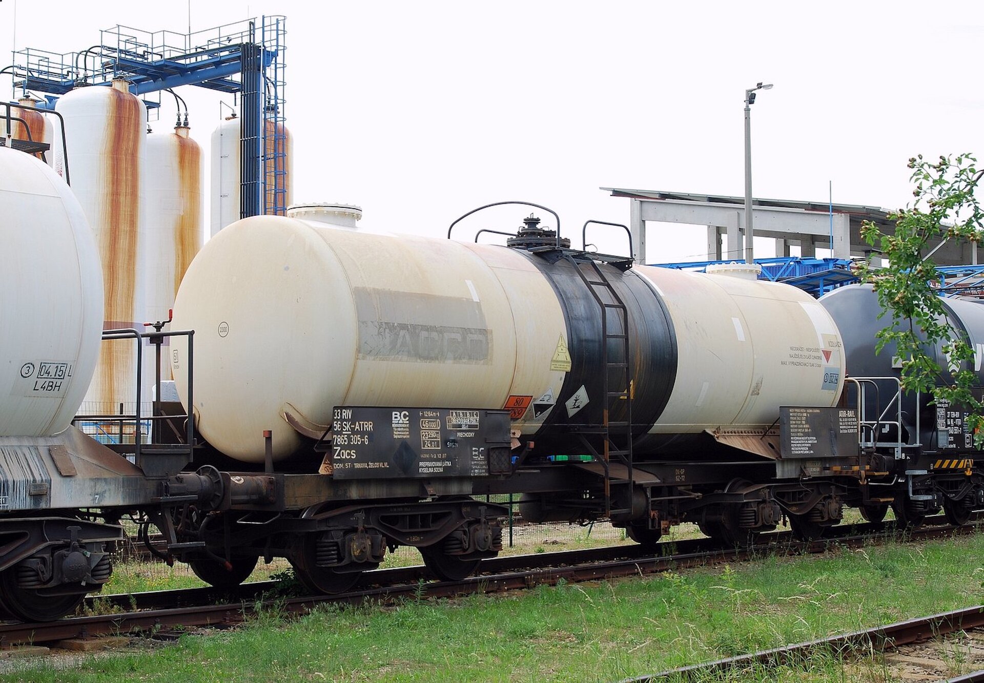 Zdjęcie przedstawia transport kolejowy z cysternami. W tle są wysokie silosy w kształcie walców. 