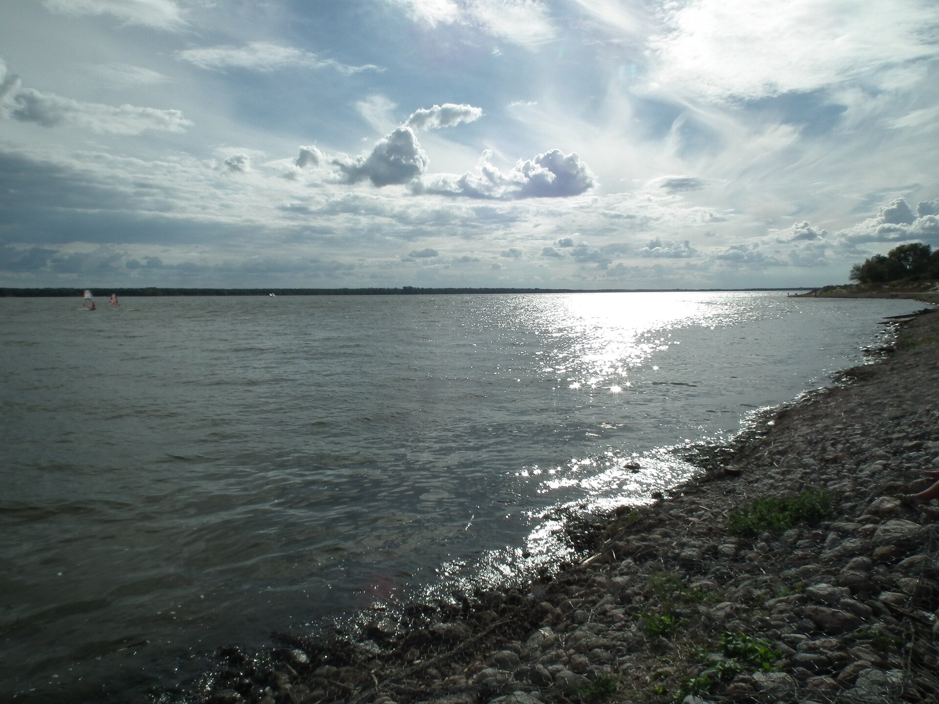 Fotografia przedstawia jezioro , po prawej stronie kamienisty brzeg, w oddali drzewa.