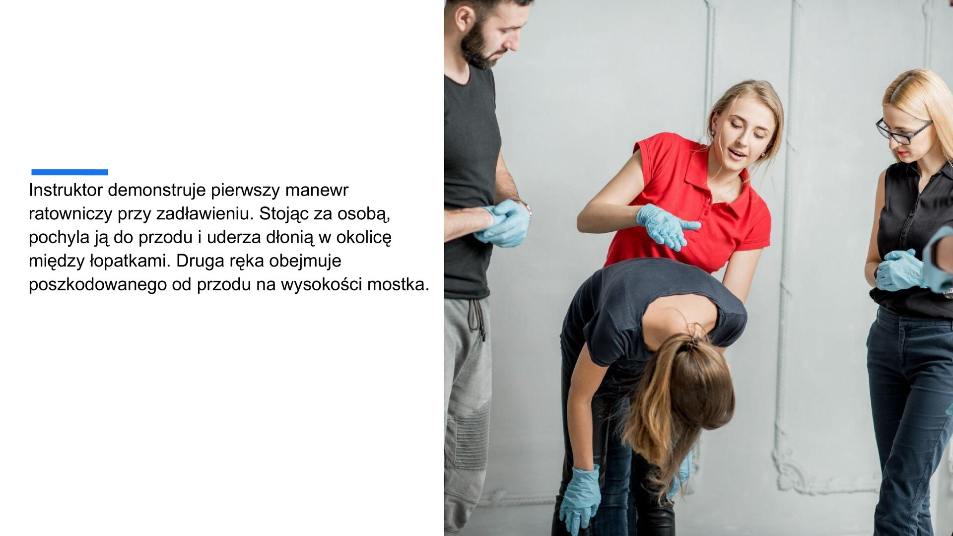 Biały slajd. Z lewej strony tekst: „Instruktor demonstruje pierwszy manewr ratowniczy przy zadławieniu. Stojąc za osobą, pochyla ją do przodu i uderza dłonią w okolicę między łopatkami. Druga ręka obejmuje poszkodowanego od przodu na wysokości mostka”. Z prawej strony zdjęcie przedstawiające cztery osoby: mężczyznę i trzy kobiety. Wszyscy mają na sobie niebieskie, silikonowe rękawiczki jednorazowe. Na środku jedna kobiet zgina się w pasie, a stojąca za nią druga kobieta, ubrana w czerwoną bluzkę, unosi prawą dłoń nad plecami drugiej kobiety.