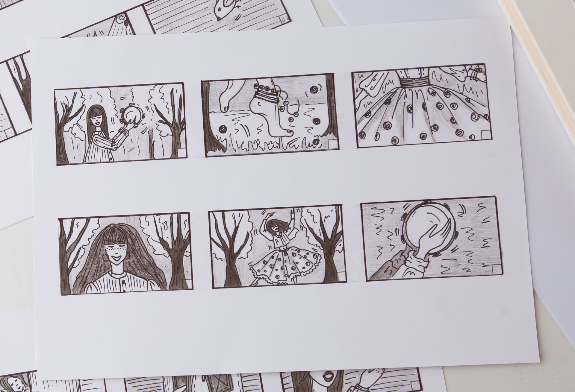 Zdjęcie przedstawiające kartkę papieru z narysowanymi sześcioma poziomymi prostokątami w środku. W każdym prostokącie znajduje się inny rysunek: dziewczynka grająca na tamburynie w lesie (plan amerykański), zbliżenie na bose stopy z koralikami wokół kostek, zbliżenie na spodnią część kwiecistej sukienki, półzbliżenie na twarz i ramiona uśmiechniętej dziewczynki (w tle widać drzewa), plan pełny ukazujący tańczącą dziewczynkę (w tle widać drzewa), zbliżenie na dłonie uderzające w tamburyn