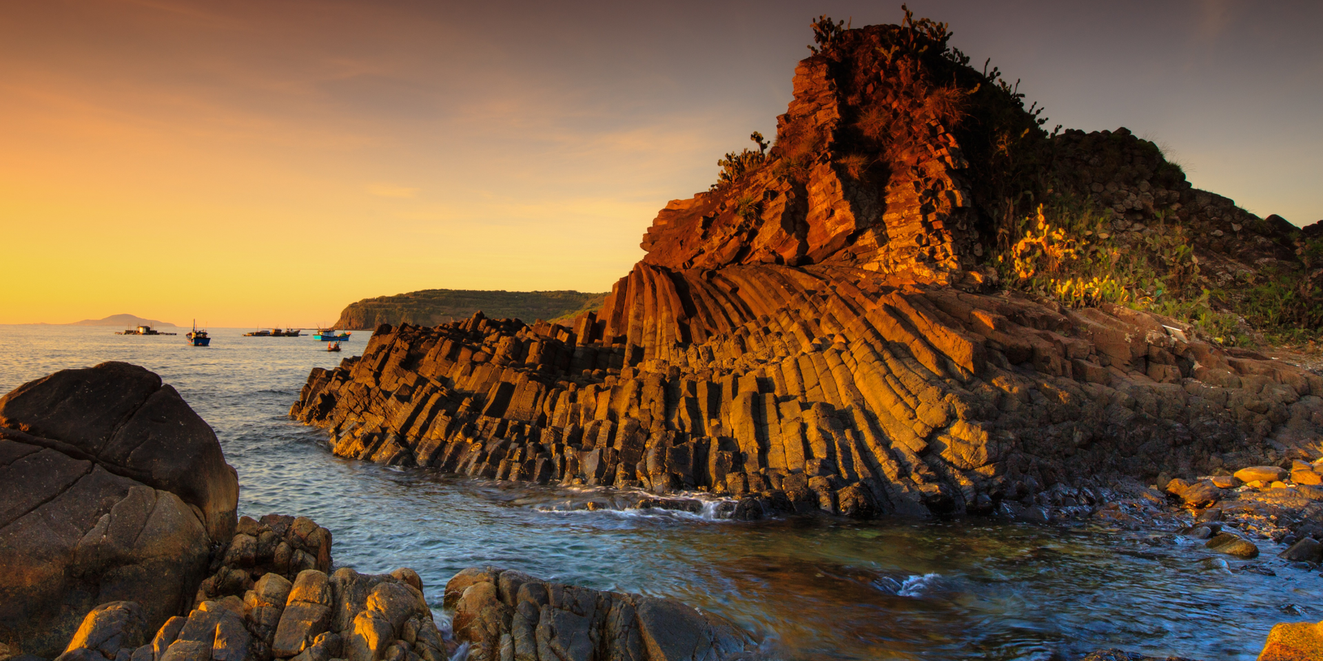 Zdjęcie przedstawia fragment wybrzeża uformowany z słupów bazaltowych. W tle znajduje się woda oraz niebo.