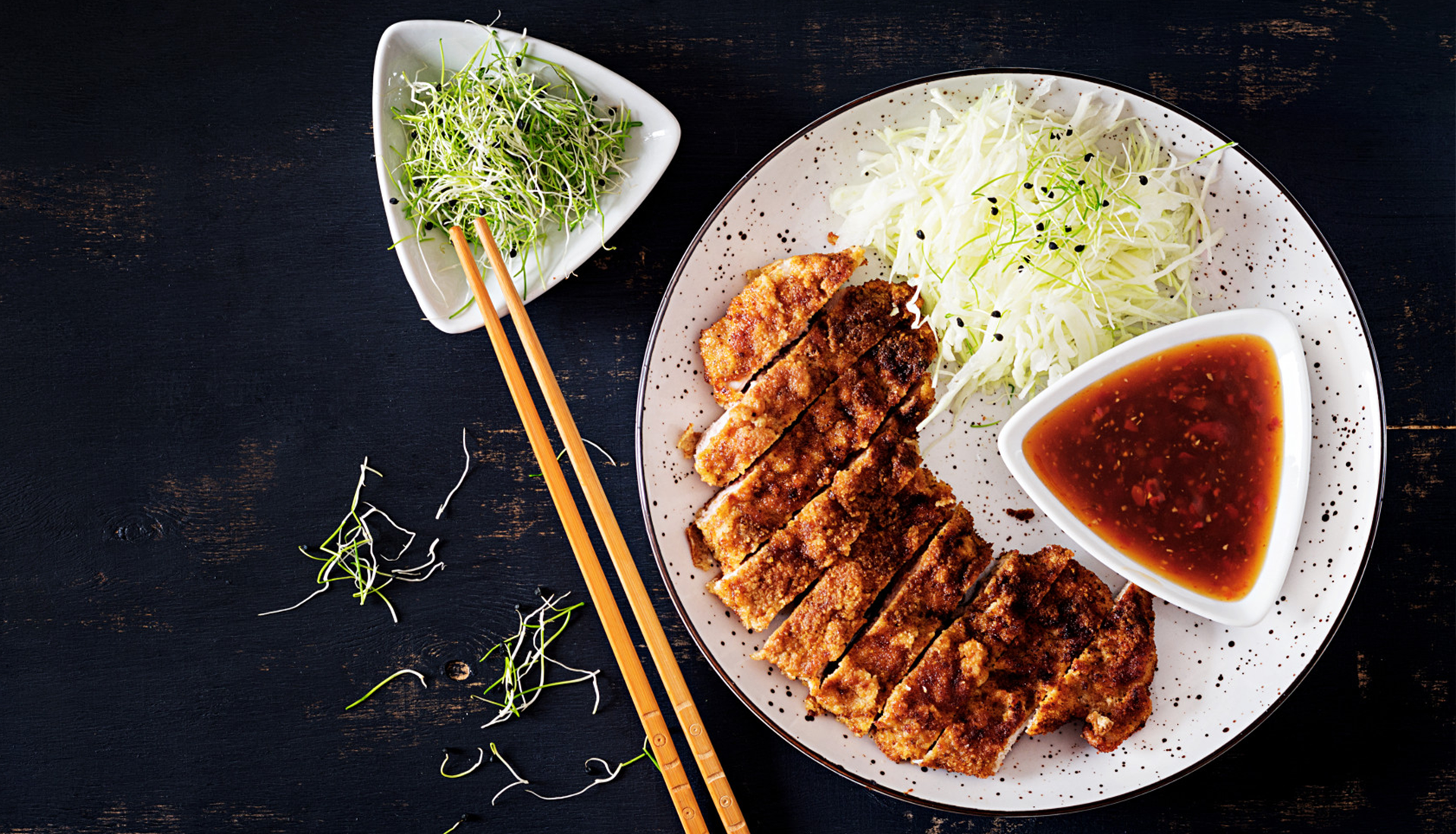 Zdjęcie przedstawia danie stojące na czarnym blacie, w ujęciu z góry. Po prawej stronie widoczny jest duży biały talerz, dekorowany ciemnymi kropkami. Na talerzu widoczna jest biała, trójkątna miseczka wypełniona czerwono‑pomarańczowym sosem, pokrojone panierowane mięso oraz surówka z poszatkowanej w cienkie paseczki białej kapusty. Na lewo od talerza, w górnej jego części, widać białą, trójkątną miseczka wypełnioną zielonymi i białymi kiełkami. O brzeg miseczki oparte są dwie drewniane pałeczki.