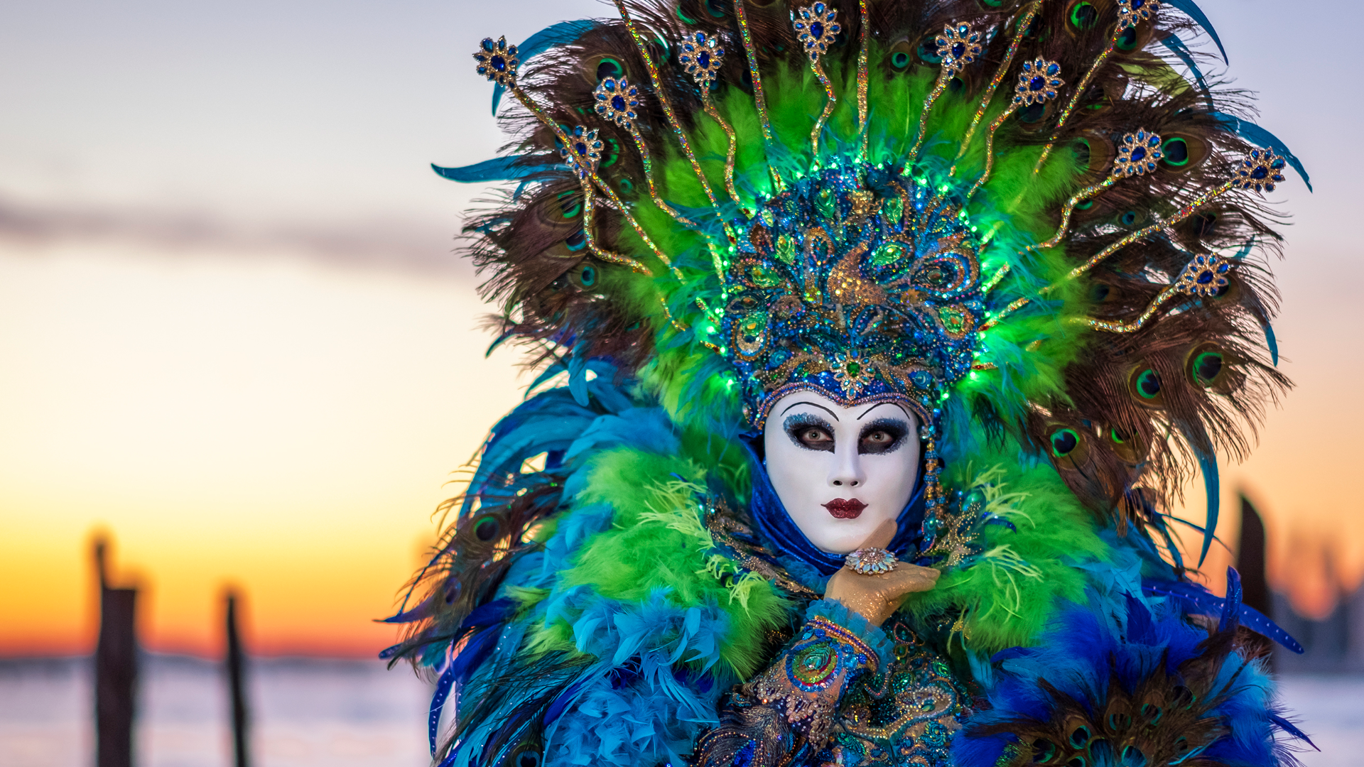 Zdjęcie przedstawia kobietę, która nosi maskę pawia. Na twarzy maska jest biała, a nad głową widoczny jest jakby rozłożony ogon pawia.