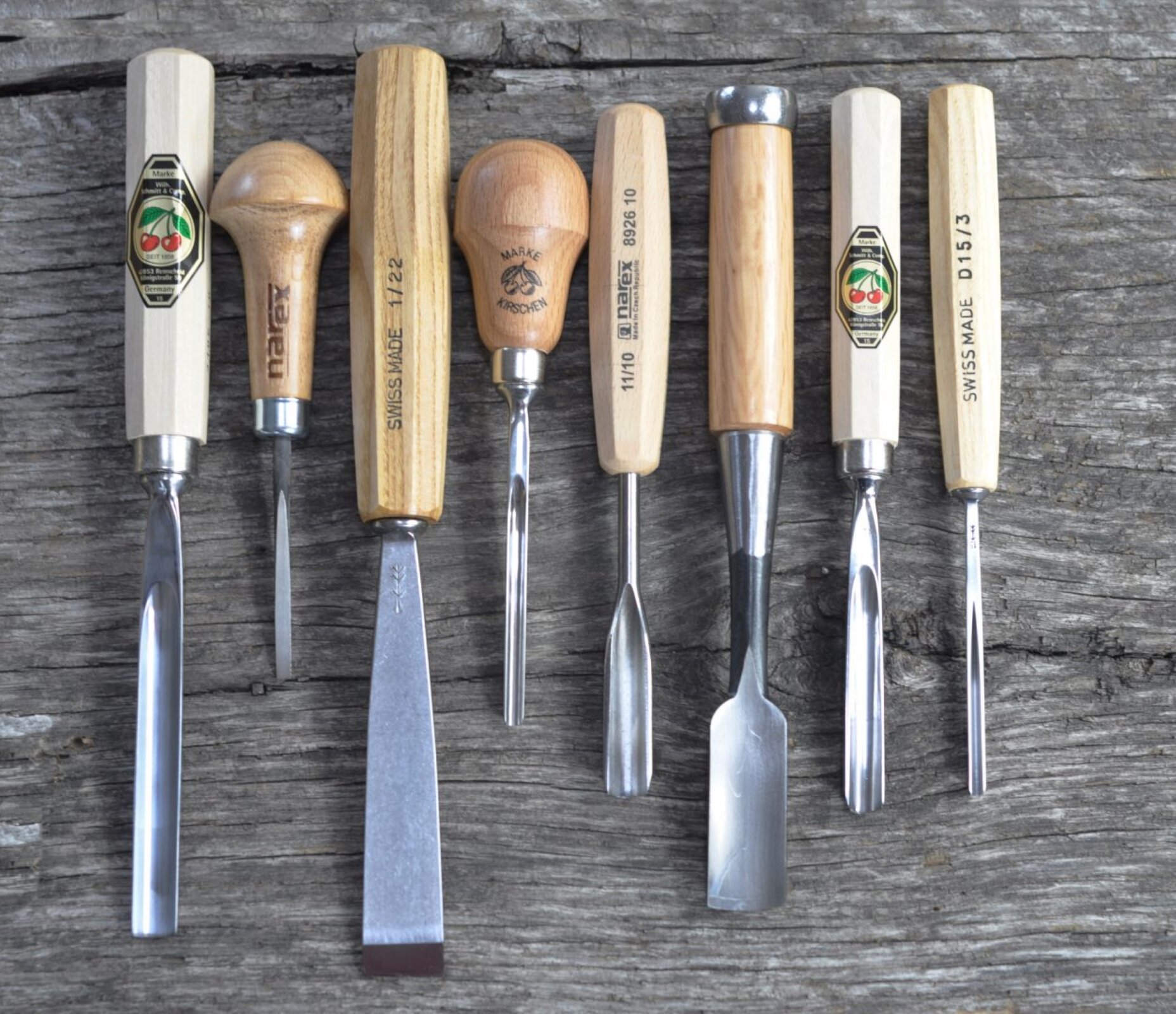 Fotografia przedstawia zestaw dłut do drewna. Każde z dłut ma brązową drewnianą rękojeść, oraz metalowe ostrze - każde dłuto ma inny kształt. Można wyróżnić dłuta o wąskim i szerokim ostrzu, okrągłe i płaskie ostrza. Dłuta leżą na drewnianym stole o szarym blacie.