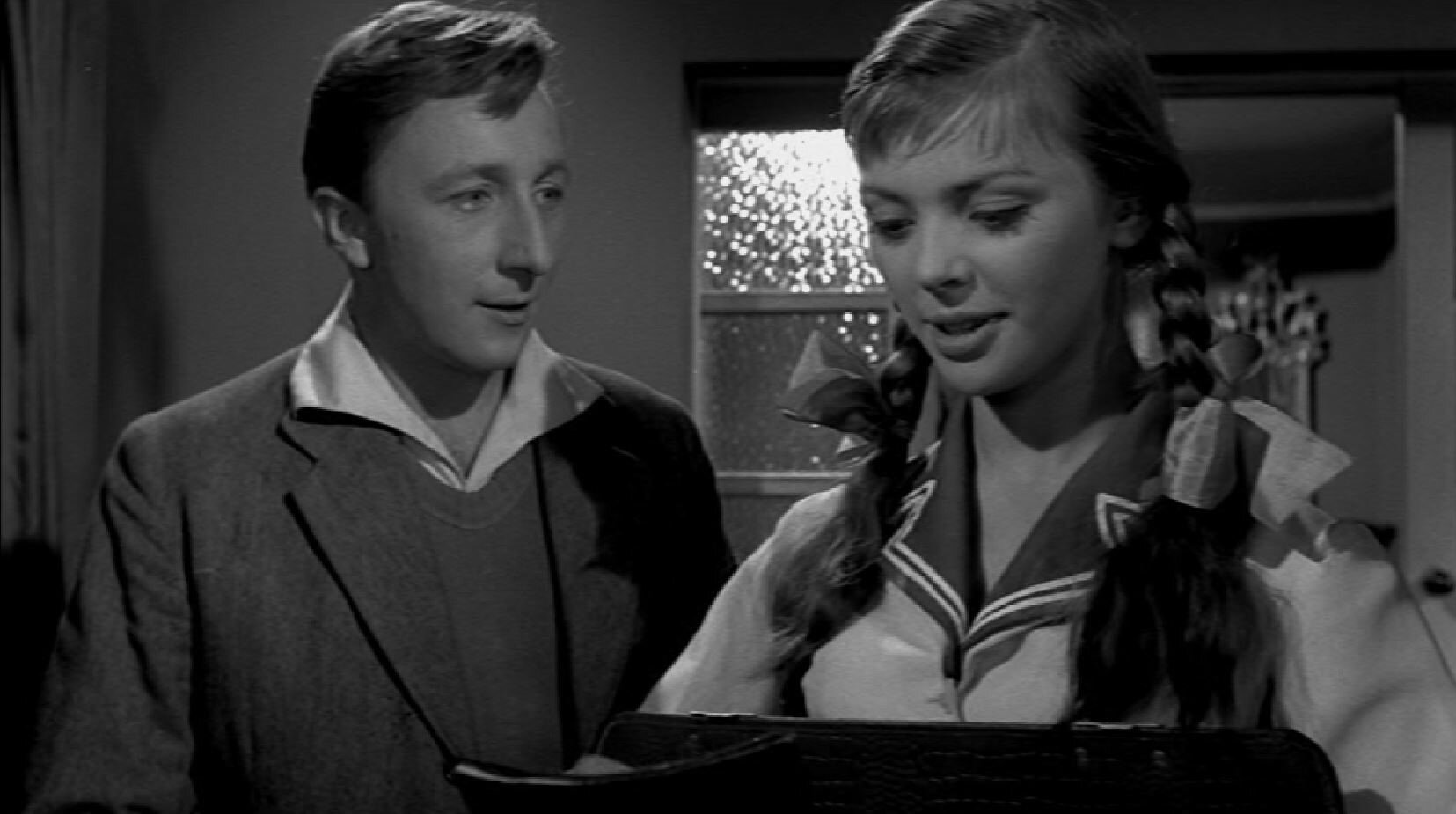 Zdjęcie przedstawia młodego mężczyznę i nastoletnią dziewczynę w mundurku szkolnym, z warkoczami i kokardami. Para rozmawia ze sobą.