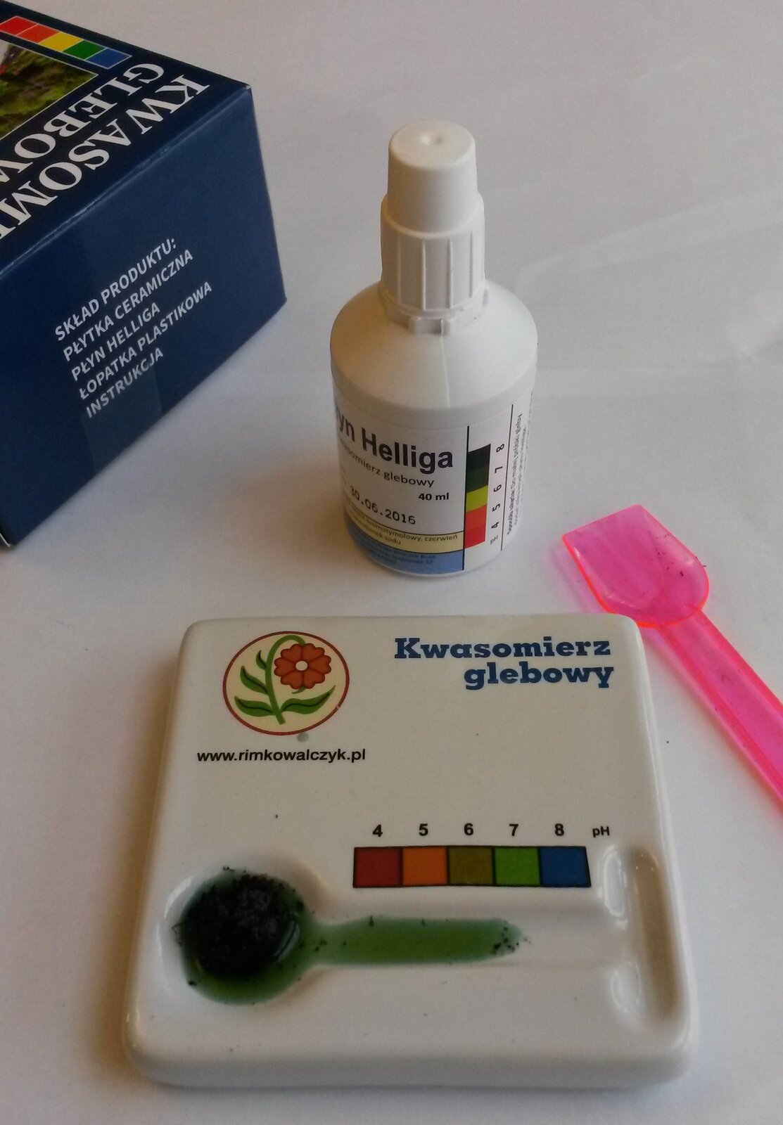Zdjęcie przedstawia zestaw do badania kwasowości gleb. Mały, plastikowy pojemnik z zakrętką, plastikowa łopatka o długości kilku centymetrów oraz plastikowa płaska płytka z okrągłym miejscem na próbkę. Obok niego pozioma skalo określająca poziom kwasowości gleby.