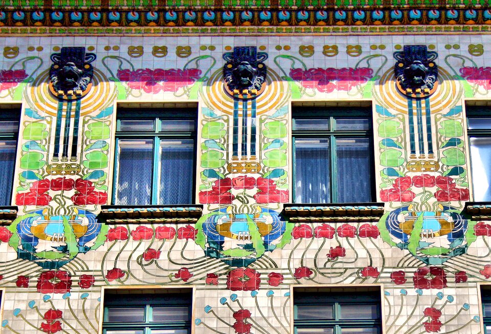 Zbliżenie fragmentu fasady wiedeńskiego Majolikahausu. Jasna elewacja pokryta jest kolorowymi płytkami ceramicznymi z powtarzającym się ornamentem czerwonych kwiatów, zielonych liści i falistych linii. Pomiędzy dekoracjami znajdują się wysokie, prostokątne okna. Nad oknami umieszczono stylizowane ciemne maski zwierzęce, z których zwisają geometryczne i roślinne ornamenty. W dolnej części widoczne są bogato zdobione parapety oraz kolejne fragmenty dekoracji kwiatowej.