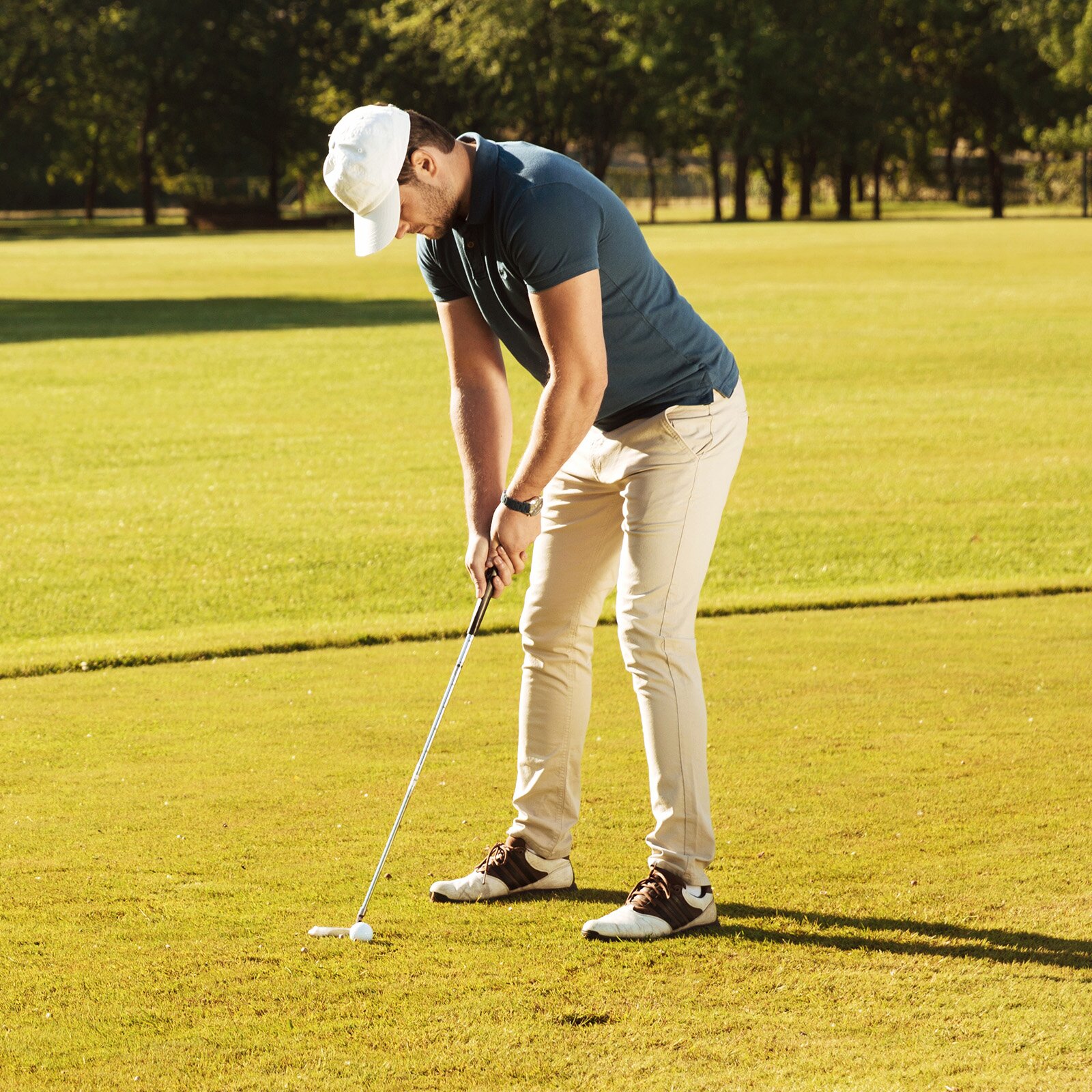 Zdjęcie przedstawia mężczyznę stojącego na przystrzyżonym trawniku, trzyma on w dłoni cienki kij zakończony stopką. Dotyka nią piłeczkę golfową. 