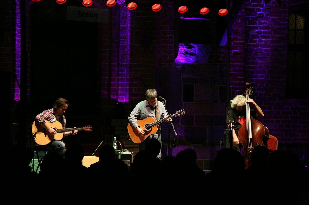 Zdjęcie przedstawia wnętrze sali koncertowej. Na scenie siedzi trzech mężczyzn. Dwóch z nich gra na klasycznych gitarach, trzeci na kontrabasie.